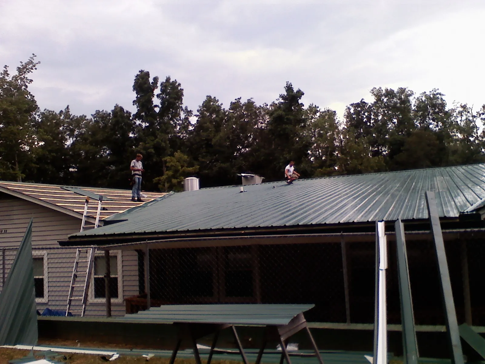 Two roofers installing green metal panels above a covered porch in Carleton